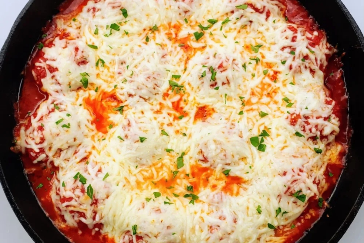 Melted cheese topping over a tomato-based dish in a cast-iron skillet, garnished with herbs.