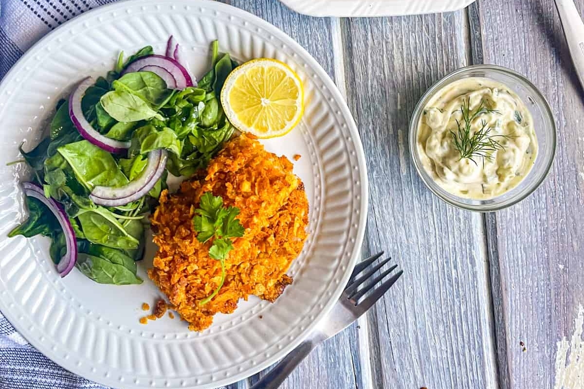 A plate of breaded fish accompanied by a side salad, lemon wedge, and tartar sauce on a wooden surface.