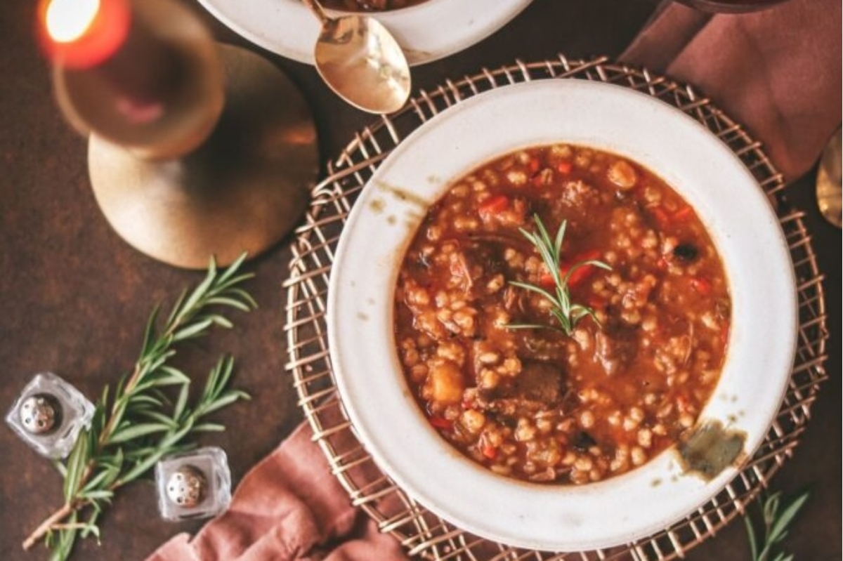 A bowl of beef and barley soup with rosemary sprigs.