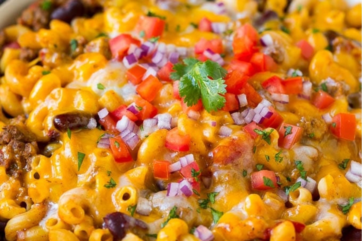 A close-up of baked macaroni and cheese topped with ground beef, melted cheddar, diced tomatoes, and onions, garnished with fresh cilantro.