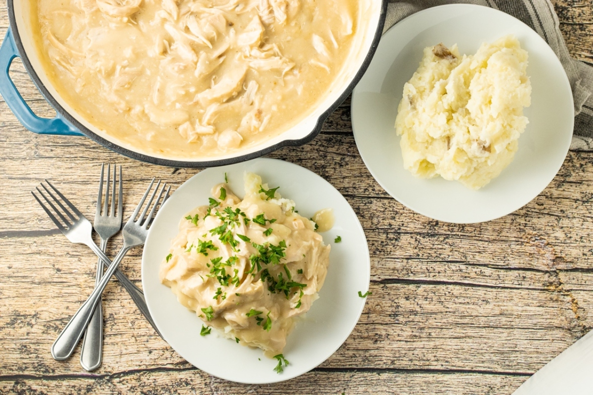 A plate of creamy chicken over mashed potatoes garnished with parsley, next to a skillet with more of the dish and a serving spoon, on a rustic wooden table.