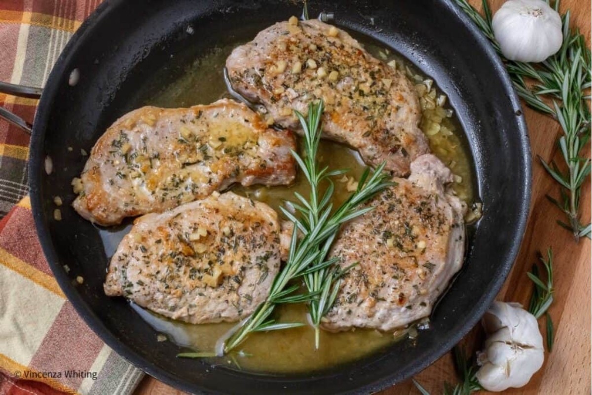 Three pork chops in a pan with garlic and rosemary, being cooked.