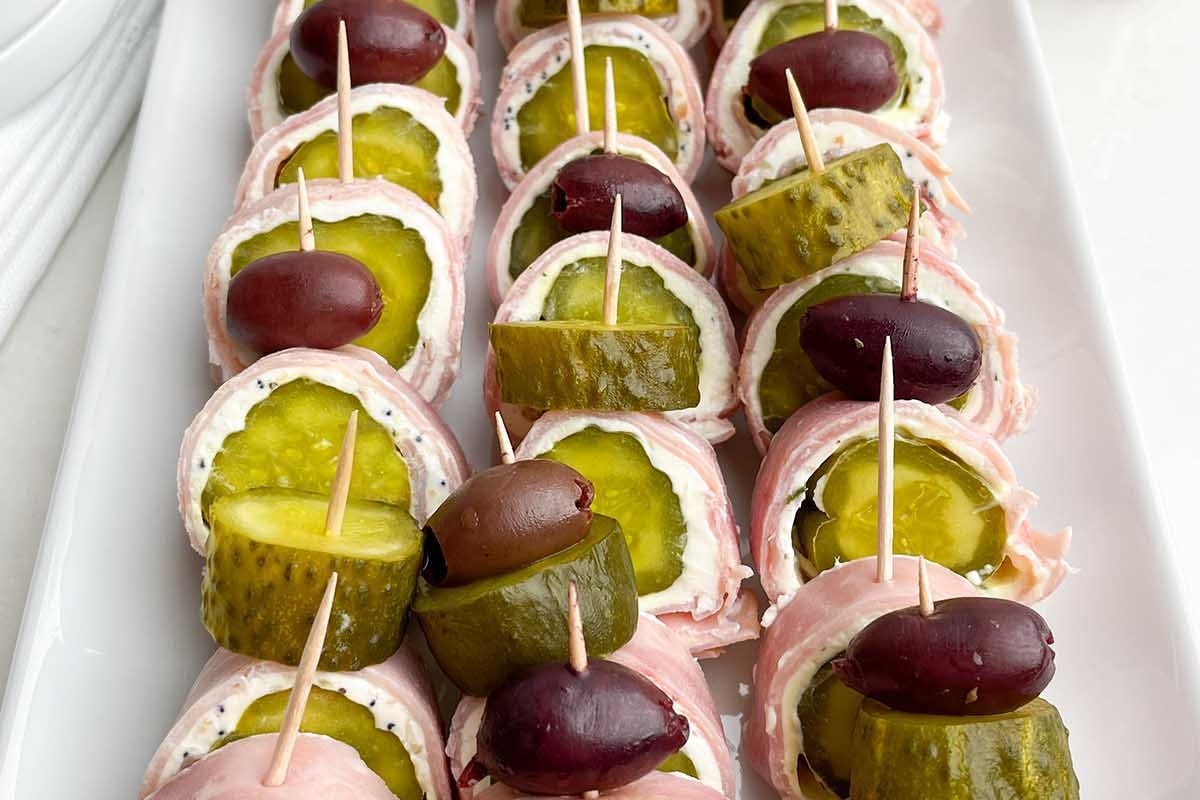 A platter of appetizers consisting of pickles and olives wrapped in slices of deli meat, secured with toothpicks.