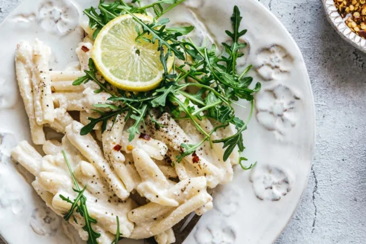 A plate of pasta with a creamy sauce, garnished with arugula and a slice of lemon.