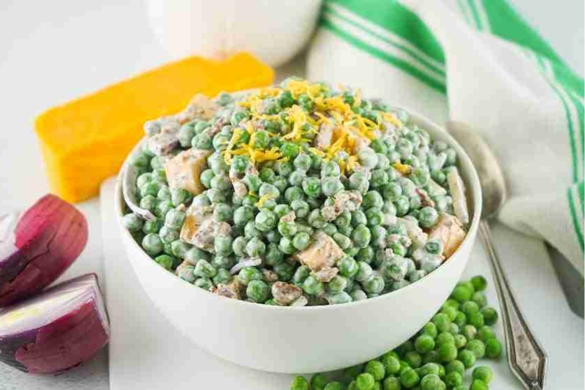 Bowl of pea salad topped with shredded cheese, with onions and cheese block in the background.