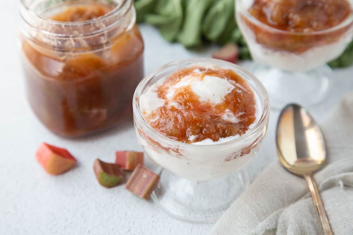 A serving of rhubarb compote with cream in a glass dish, accompanied by a spoon and additional compote in a jar, on a light surface.