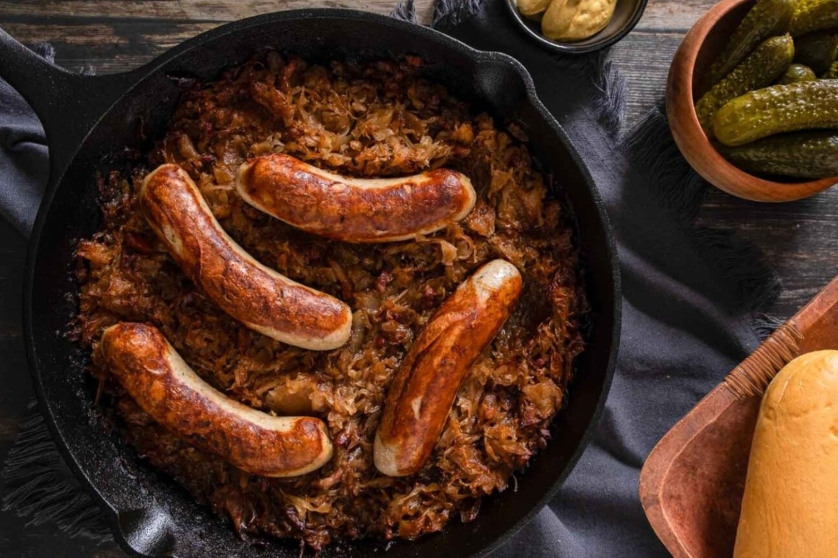 Sausages served with sauerkraut in a cast-iron skillet, accompanied by pickles and bread on a wooden table.