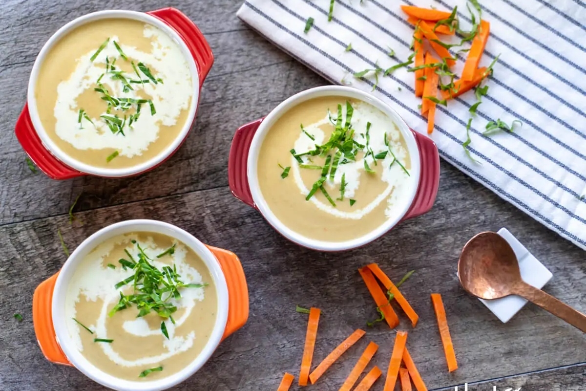 Three bowls of creamy soup garnished with herbs on a wooden table with carrot peels and a striped napkin on the side.