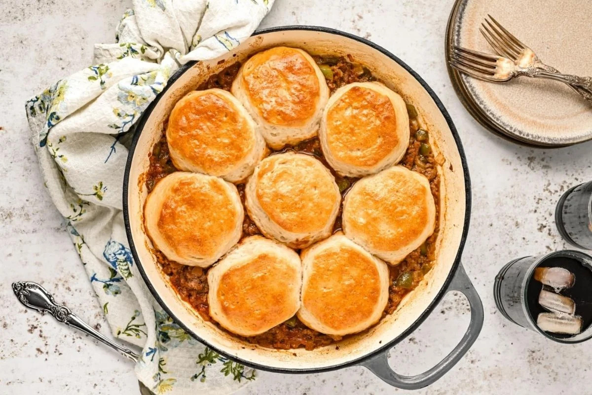 A skillet of savory cobbler with golden-brown biscuits on top, surrounded by a spoon, a cloth, and seasonings on a speckled countertop.