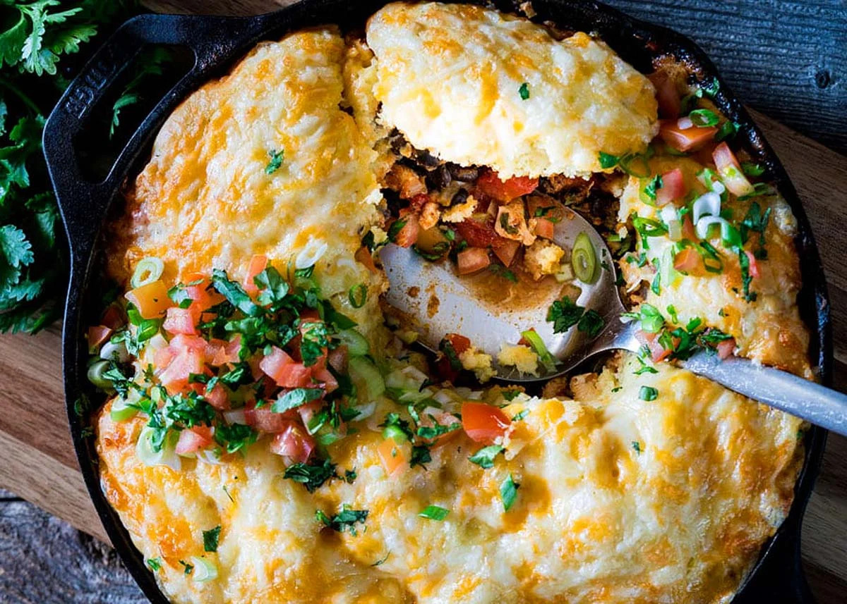 Cast iron skillet with a baked cheesy casserole topped with tomatoes and green onions, served with a spoon.