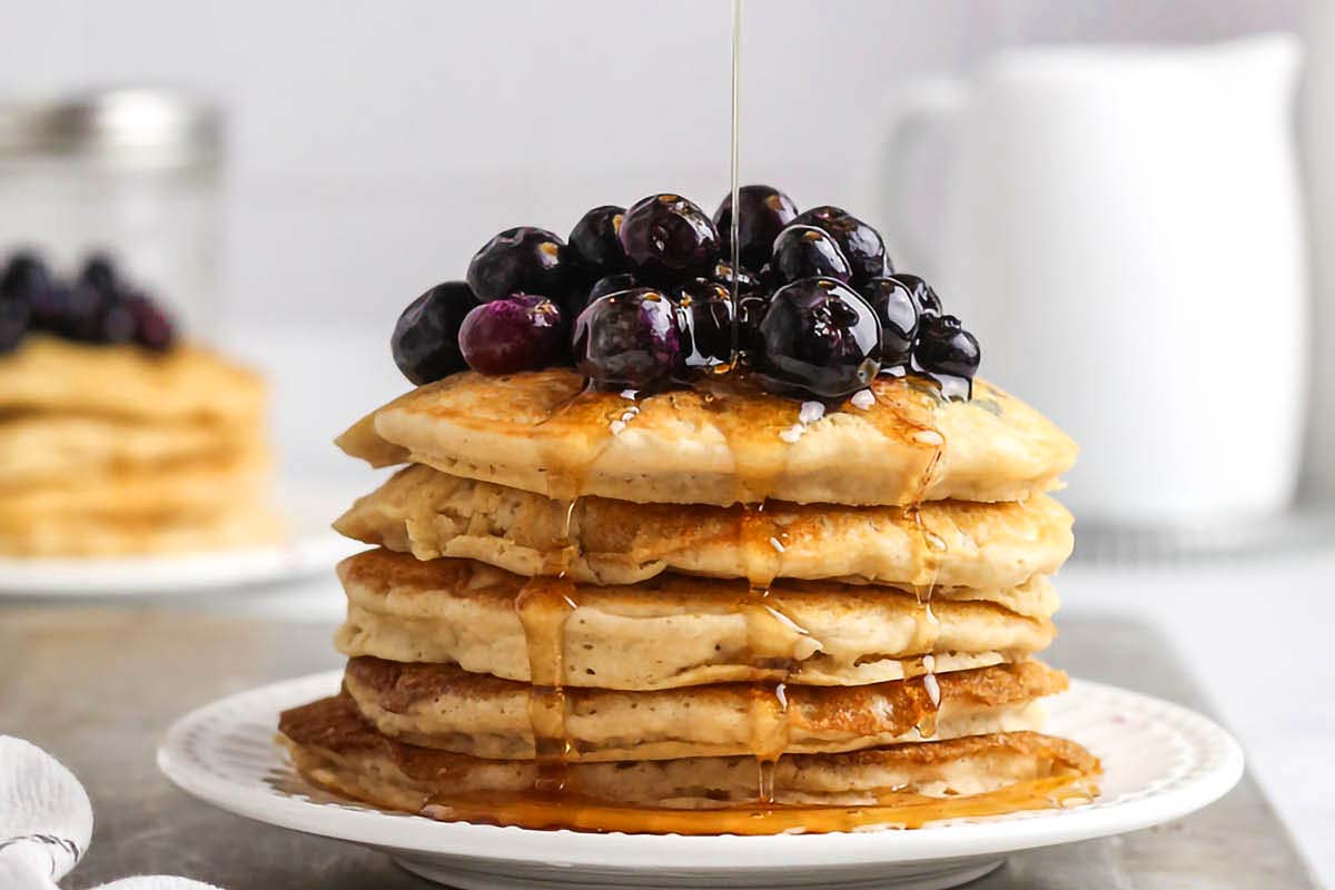 Stack of pancakes with blueberries on top and syrup being poured over, served on a white plate with a blurred background.