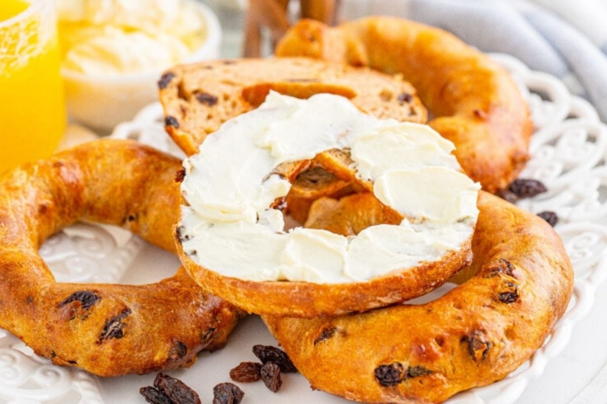 A pile of bagels on plate showing half of one with cream cheese on it.