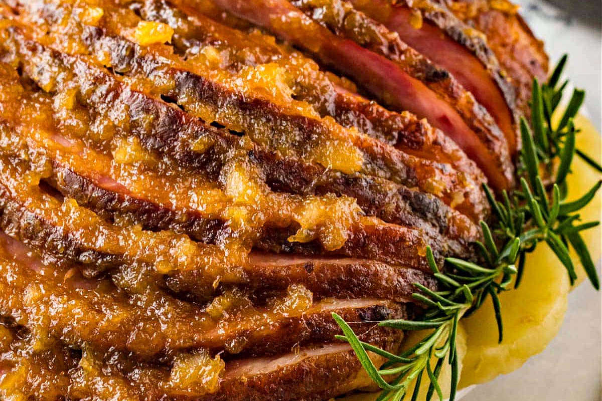 Close-up of a sliced, slow cooker glazed ham garnished with rosemary sprigs, resting on a bed of mashed potatoes.
