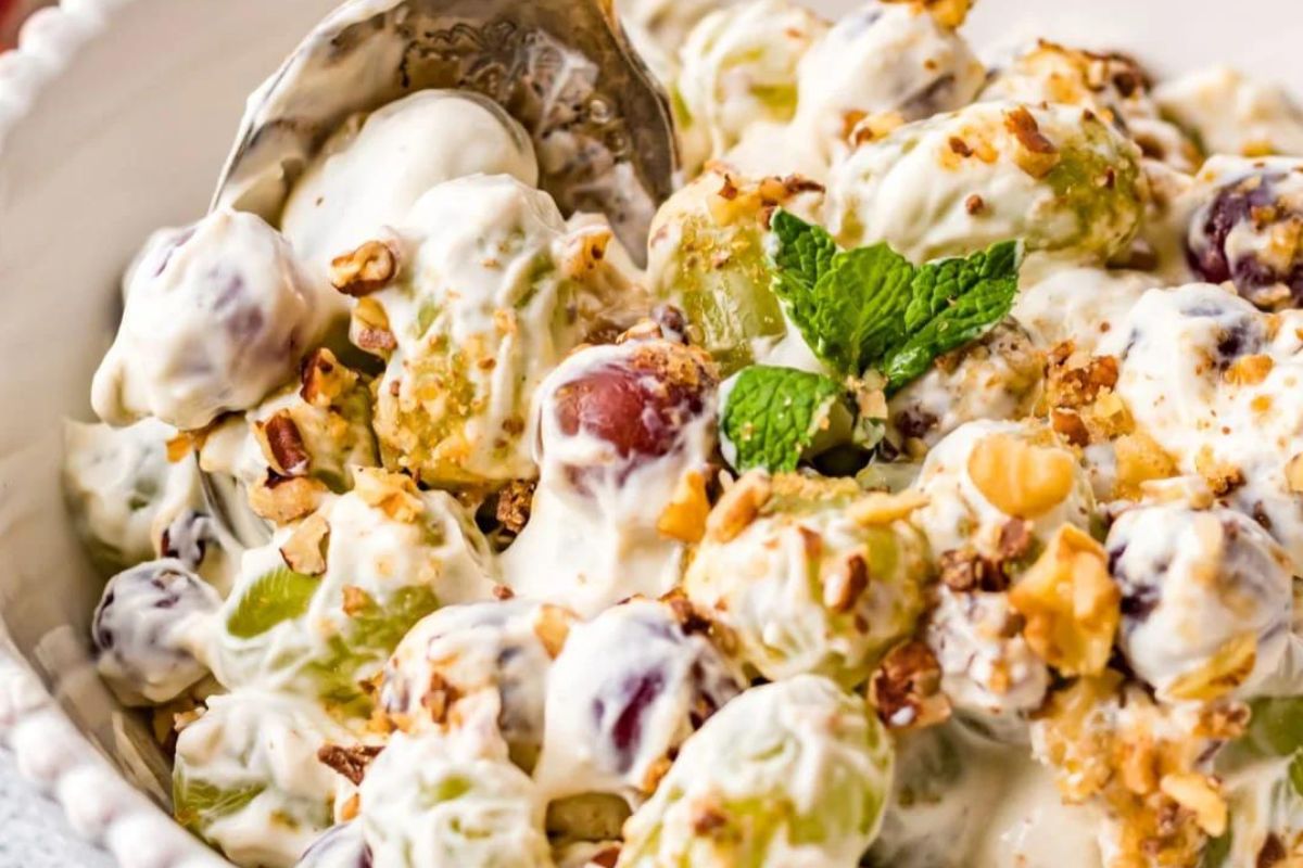 A bowl of grape salad topped with chopped nuts and garnished with a mint leaf, being served with a spoon.