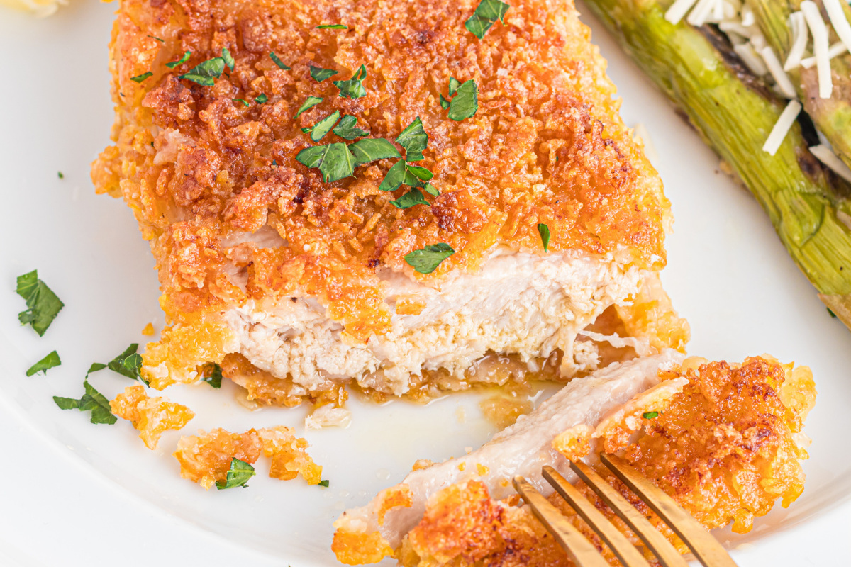 Close-up of a fork cutting into a crispy, golden-brown breaded chicken breast garnished with chopped parsley. Grilled asparagus with grated cheese is visible in the background on the plate.