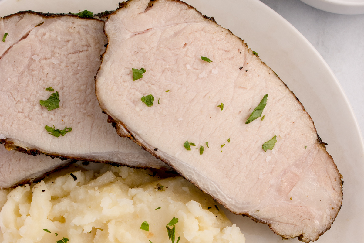 Two slices of irresistible pork garnished with herbs are served with mashed potatoes, all presented on a white plate.