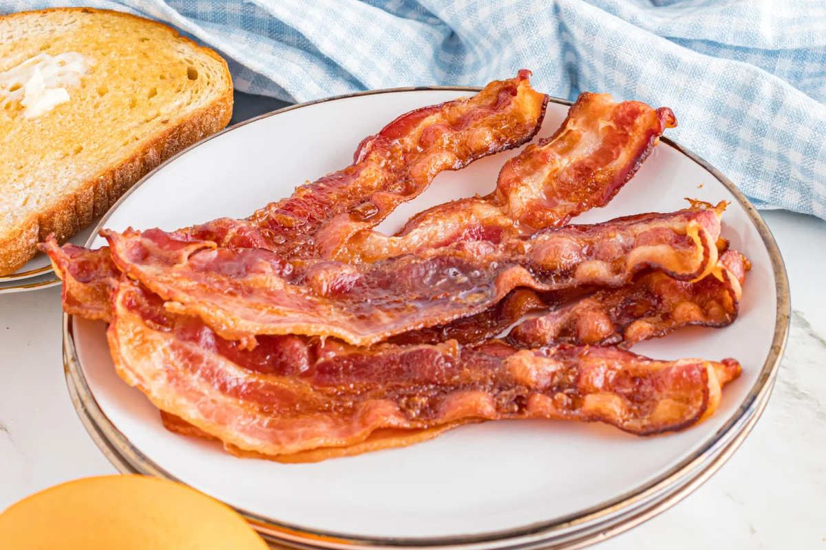 A plate with several strips of cooked bacon next to a slice of buttered toast sits on a table with a blue checkered cloth, showcasing one of those irresistible recipes perfect for both breakfast and brunch.