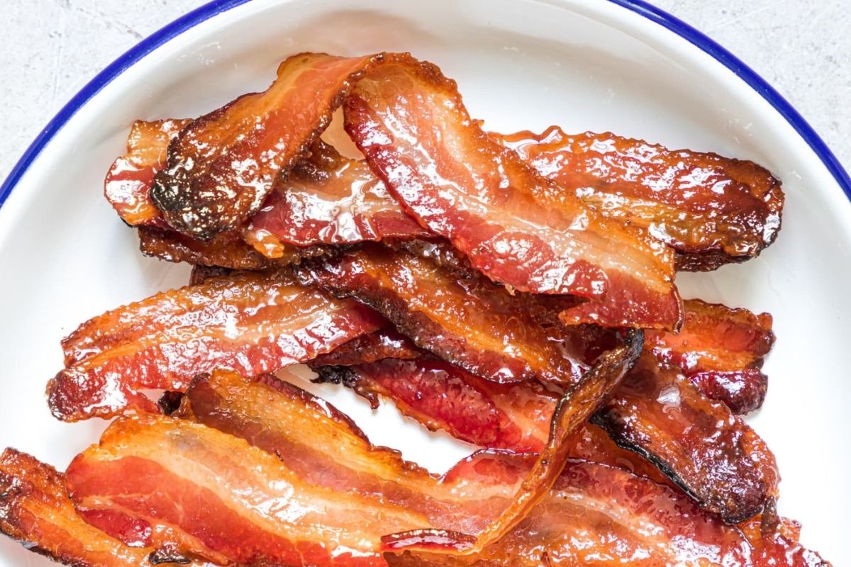 A plate of cooked bacon strips, with a crispy, caramelized appearance. The plate has a blue-rimmed edge.