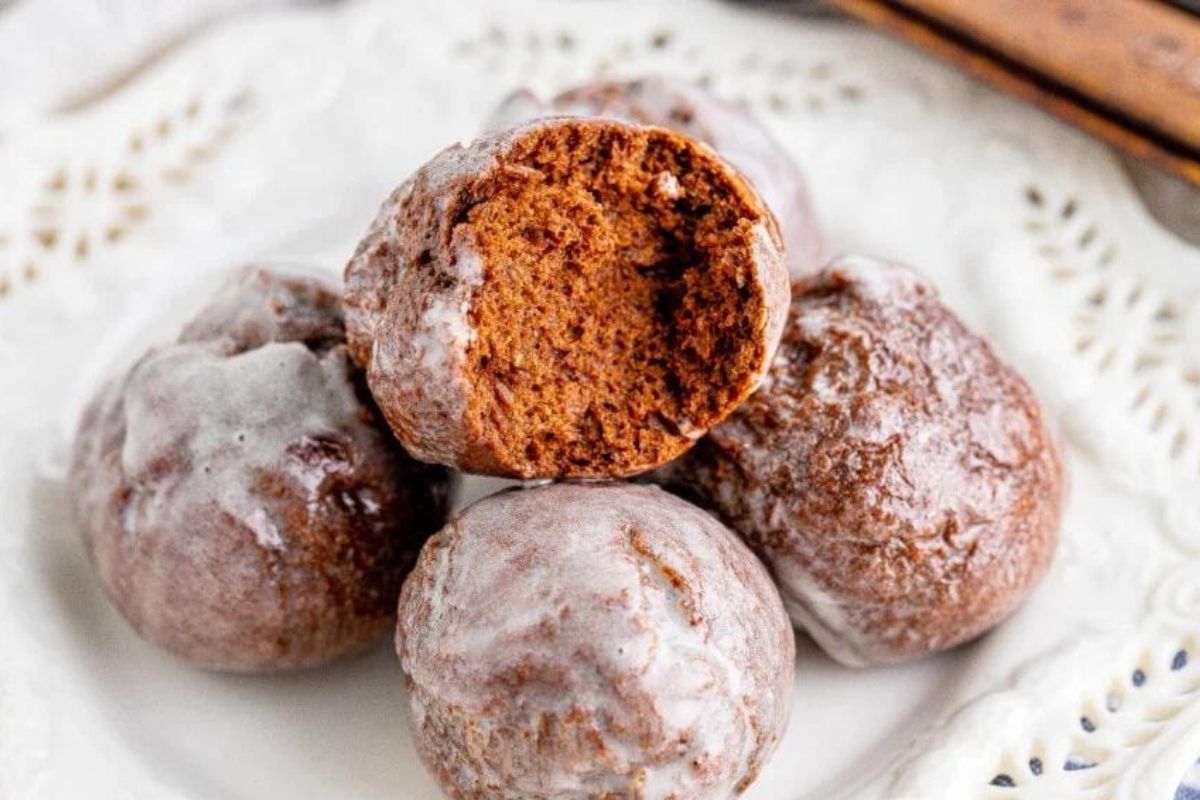 Four glazed chocolate cookies are stacked on a white plate. One cookie has a bite taken out, revealing a soft, brown interior.