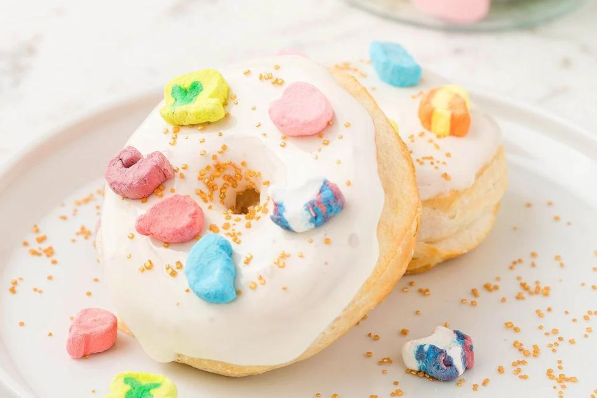 Two doughnuts with white icing, topped with colorful cereal marshmallows and golden sprinkles, placed on a white plate.
