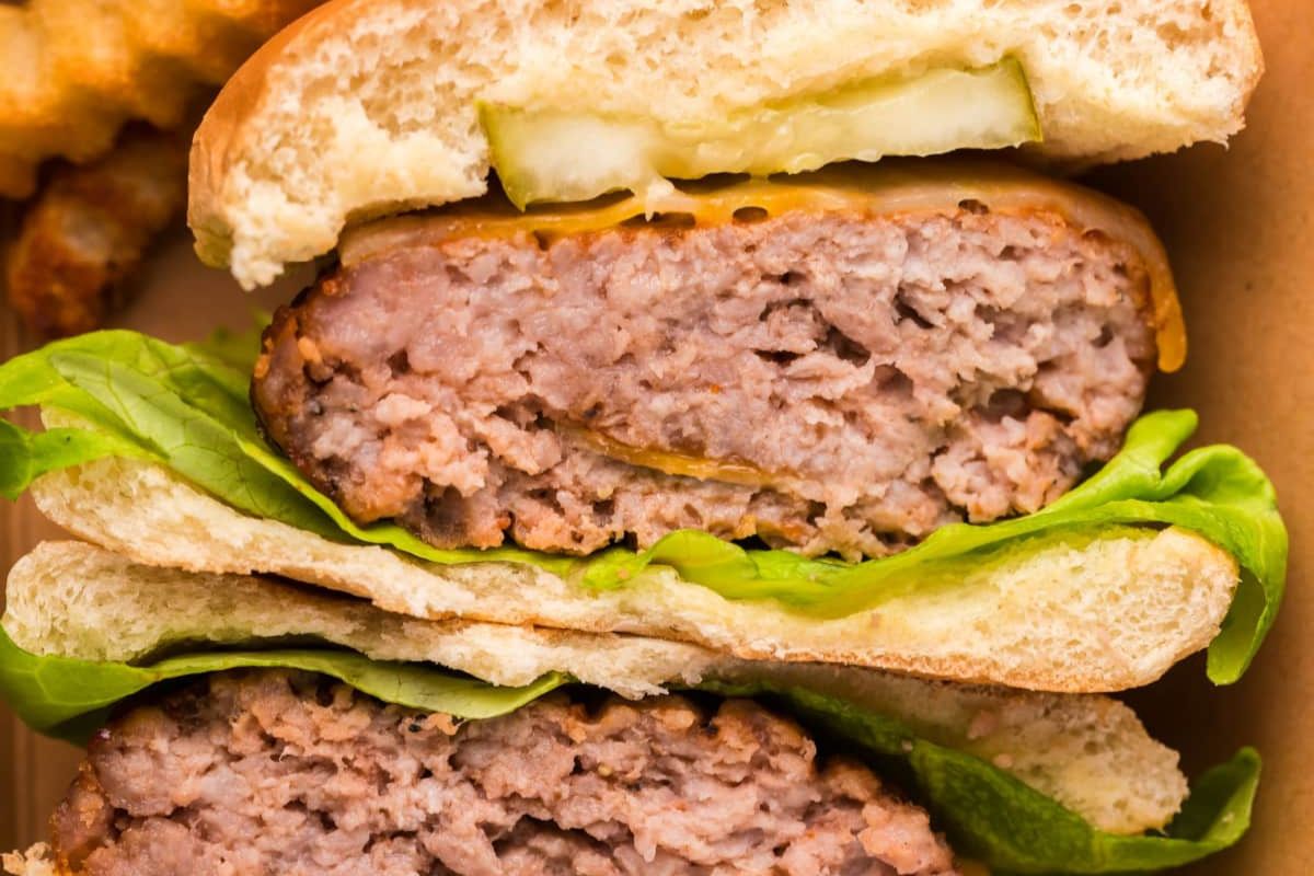 Close-up of a halved hamburger showing layers of lettuce, beef patty, cheese, and pickles between slices of a bun, with crinkle-cut fries in the background.