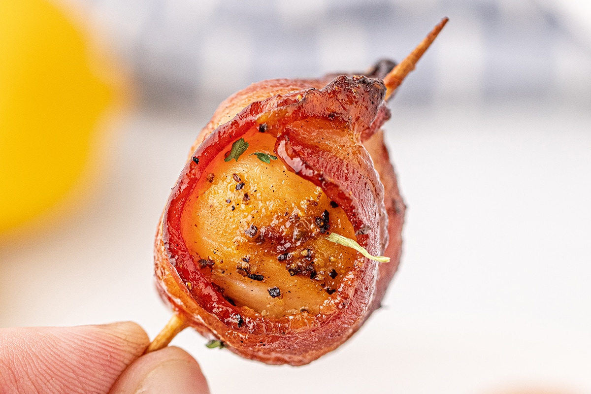 A close-up of a bacon-wrapped scallop on a toothpick, seasoned with herbs and black pepper, held by a hand against a blurred background. This smoked dinner delight is an absolute must-try for any occasion.