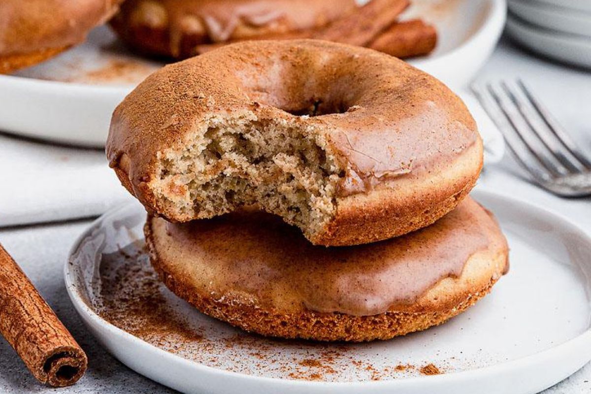 Two cinnamon glazed donuts are on white plates, one with a bite taken out. There is a cinnamon stick and a fork in the background.