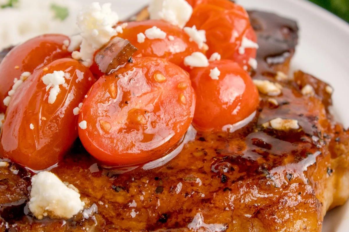 A close-up view of a grilled steak topped with cooked cherry tomatoes and crumbled feta cheese.