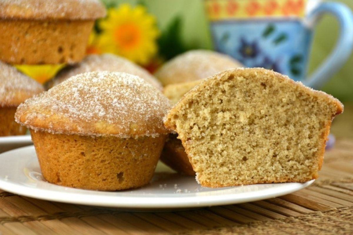A plate with four sugar-topped muffins, one of which is cut in half, showcasing its interior crumb. In the background, there is a floral-patterned mug and a blurred flower arrangement.