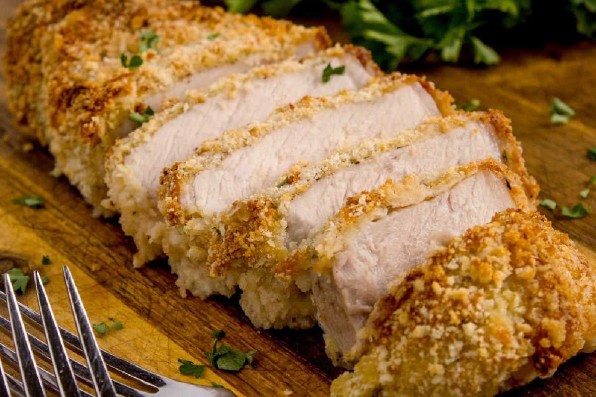 Sliced, breaded pork chop on a wooden cutting board, garnished with parsley, with a fork on the side.