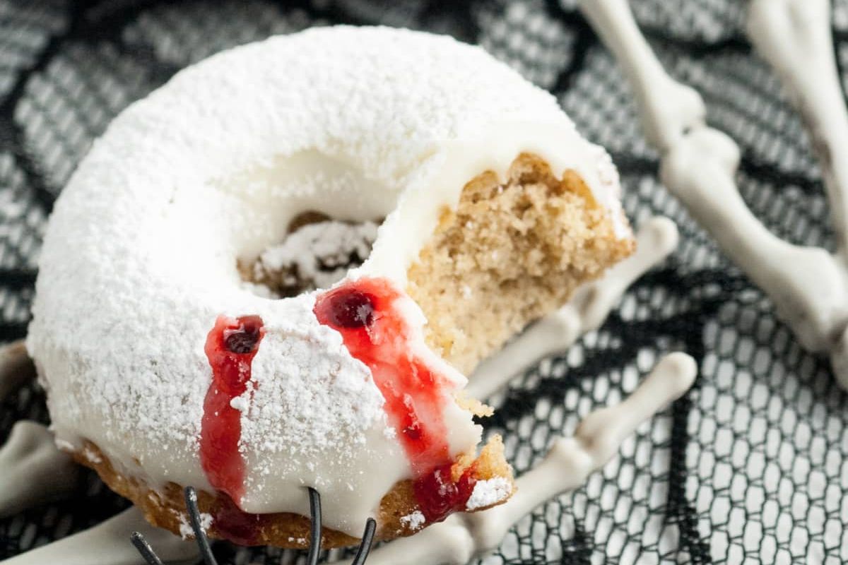 A bitten doughnut topped with white icing and red syrup sits on a black lace surface, surrounded by plastic skeleton hands.