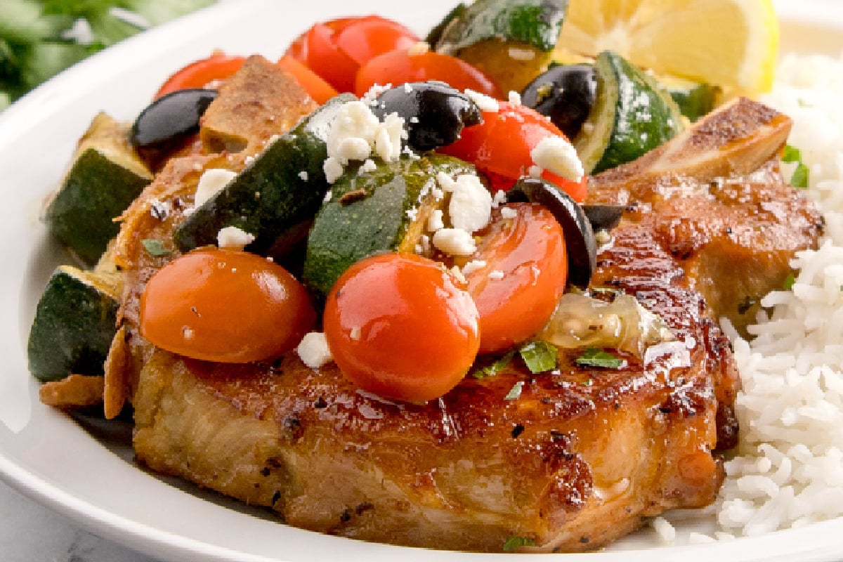 Grilled pork chop served with rice, topped with cherry tomatoes, zucchini, black olives, and crumbled feta, with a lemon wedge on the side. This dish is one of our