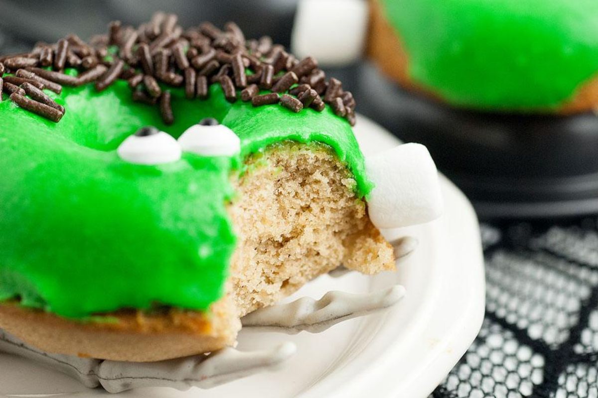 A doughnut with green icing, chocolate sprinkles, and candy eyes is partially eaten. It is placed on a white plate, and appears to be decorated to look like a frog.