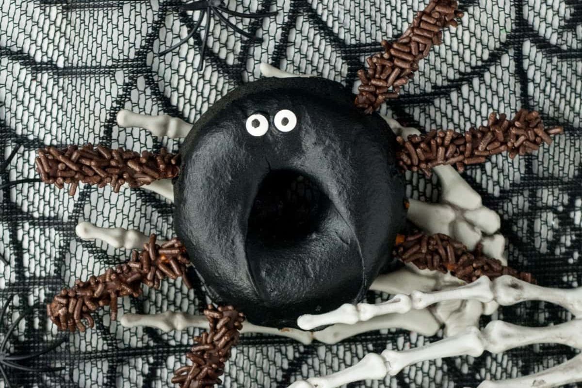 A black donut with two small white eyes is decorated with chocolate sprinkles to resemble a spider, placed on a web-patterned background with skeleton hand props.