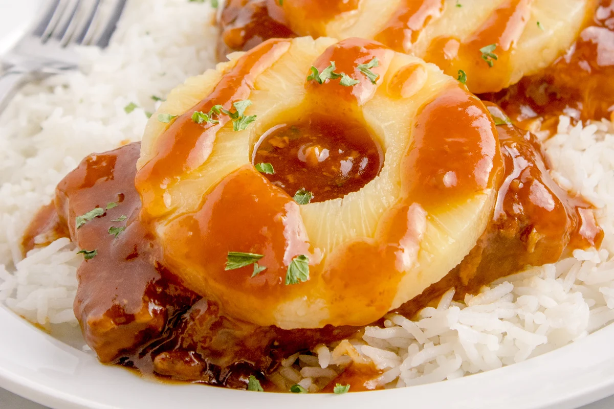 A plate of white rice topped with a grilled pork chop, covered in a tangy sauce and garnished with a pineapple ring and chopped parsley.