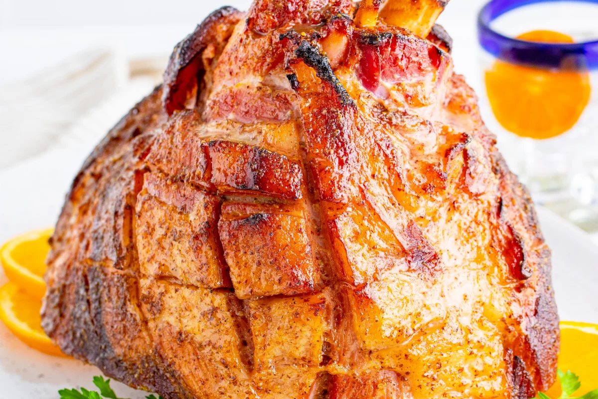 A close-up of a glazed, roasted ham garnished with orange slices and herbs, with an orange-garnished drink in the background.