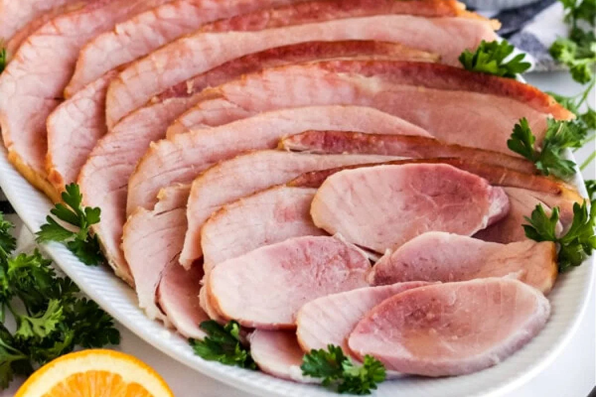 A platter of thinly sliced ham, from our irresistible pork recipes collection, garnished with parsley and complemented by an orange slice on the side.