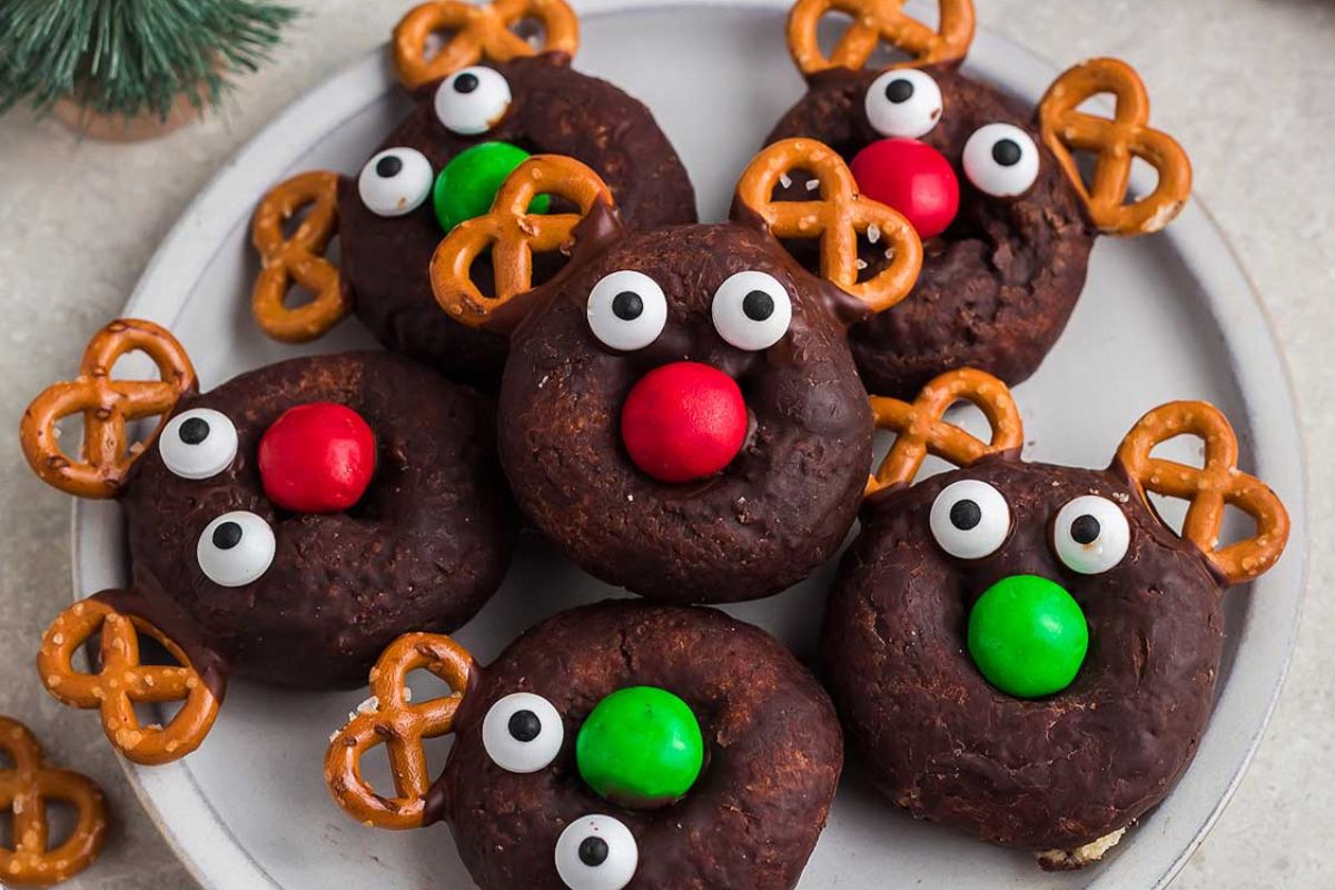 Homemade reindeer donuts with pretzel antlers, candy eyes, and colorful candy noses on a white plate.