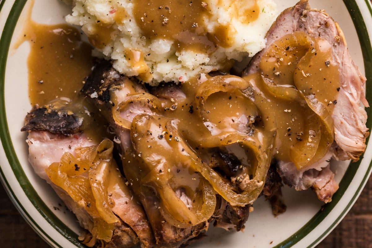 Plate of roasted meat topped with gravy and sliced onions, served with mashed potatoes and more gravy.