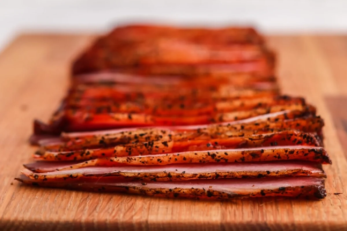 Sliced pieces of seasoned bacon are neatly arranged on a wooden cutting board, making it a must-try complement for smoked dinners.