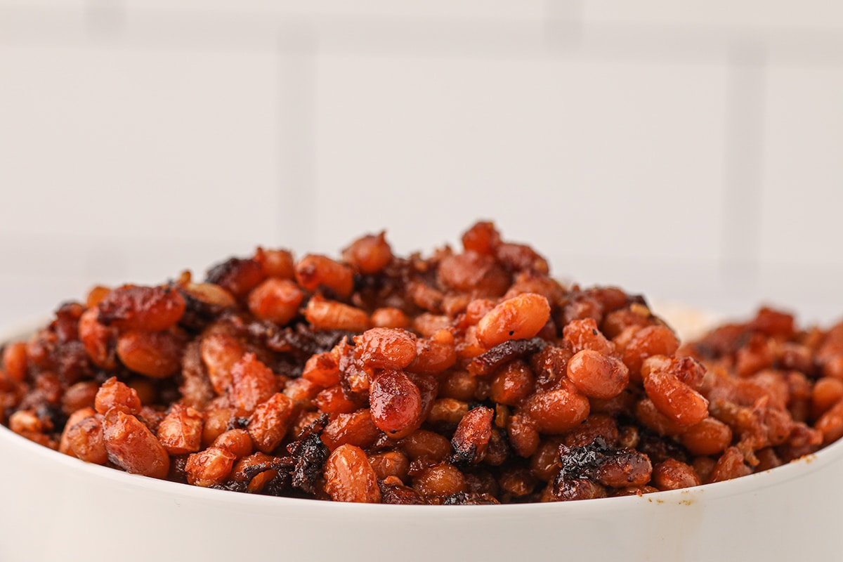 A close-up of a bowl filled with cooked pinto beans mixed with chunks of smoked bacon, set against a soft white background.