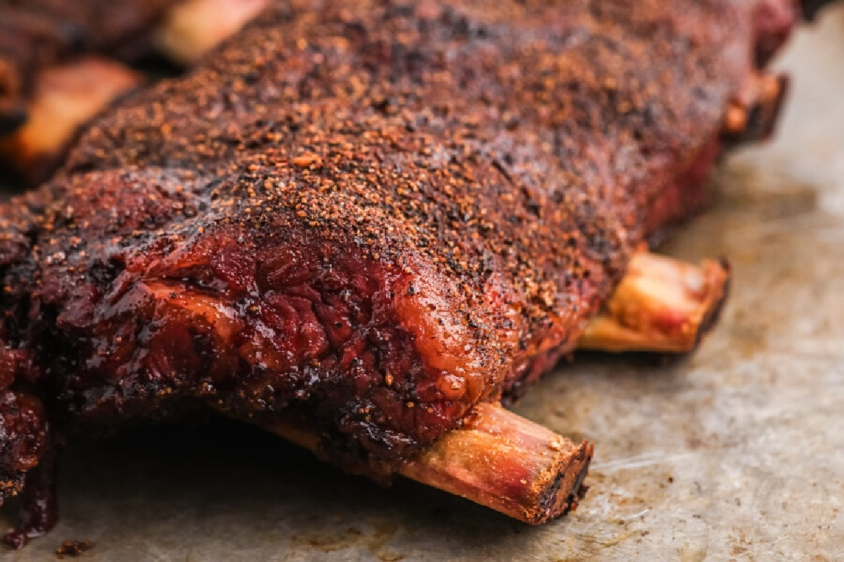 Close-up of a rack of cooked, seasoned beef ribs, drawing inspiration from smoked recipes, placed on a gray surface.