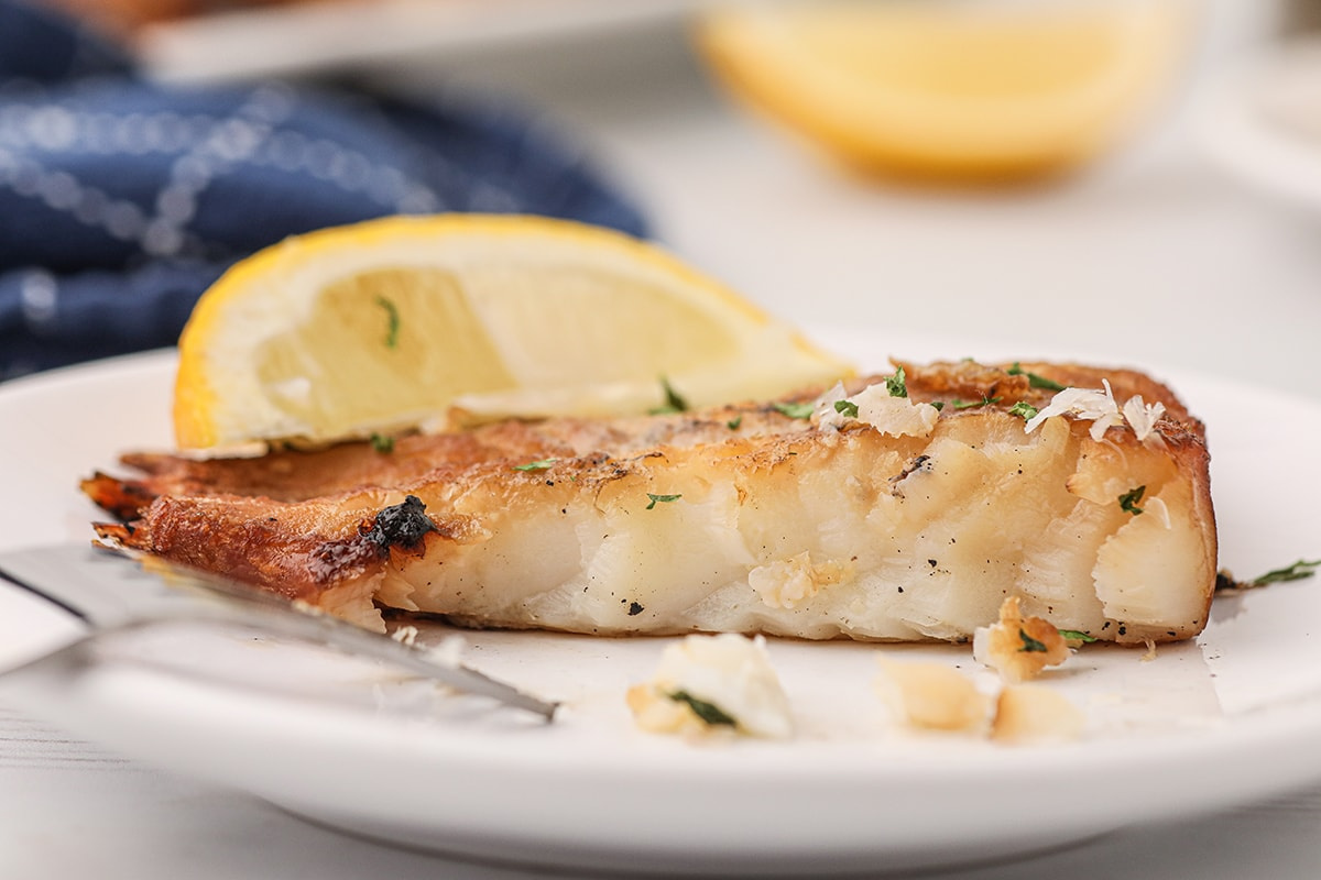 Close-up of a grilled white fish fillet garnished with herbs, served on a white plate with a lemon wedge and a fork, capturing the essence of smoker recipes in every savory bite.
