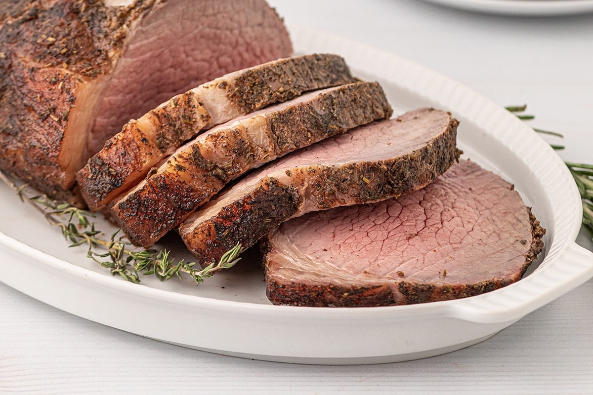 A cooked roast beef with three slices cut off, garnished with rosemary sprigs, presented on a white dish, is perfect for smoked dinners—a must-try delight complete with delectable sides.