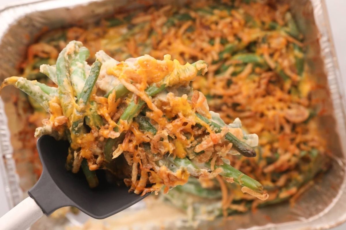 A spatula holds up a serving of green bean casserole, featuring crispy onions and cheese, above a round aluminum tray of the dish.