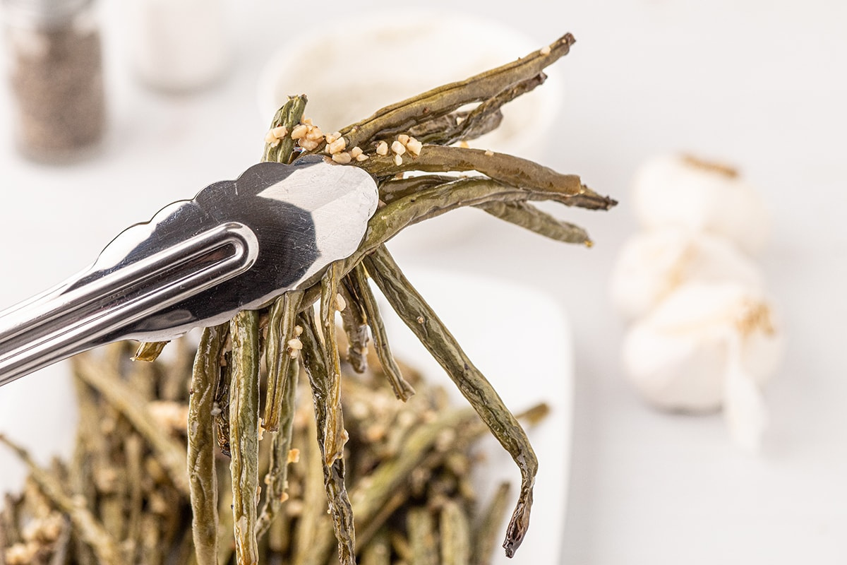 Tongs holding smoked green beans sprinkled with sesame seeds, with garlic and condiments blurred in the background.