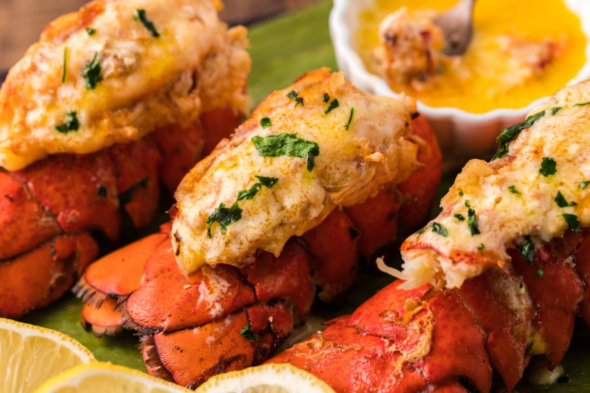 Close-up of baked lobster tails garnished with parsley, served with a side of melted butter in a small dish, and lemon wedges on the side, perfect for those exploring smoker recipes.