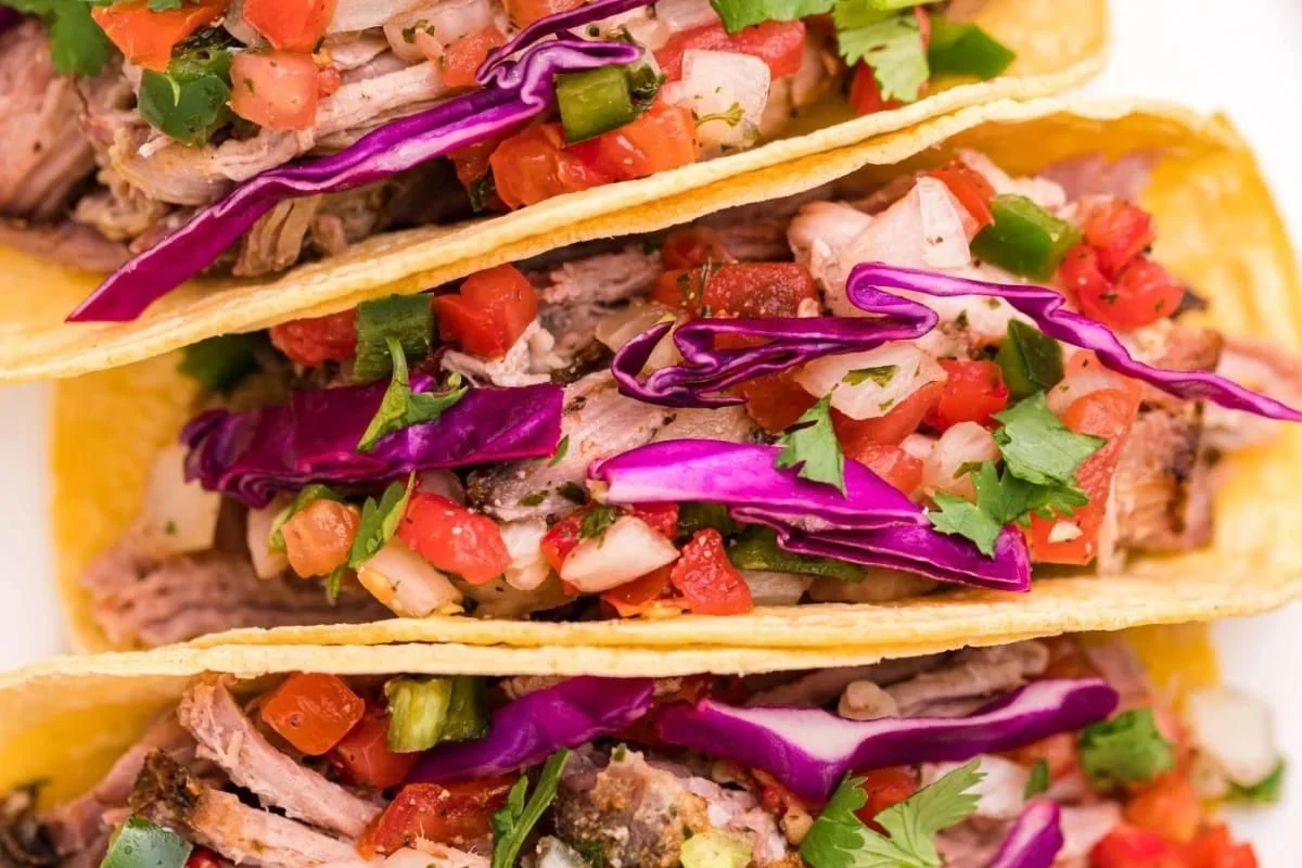 Three tacos filled with smoked shredded meat, diced tomatoes, red cabbage, and cilantro in crispy yellow corn shells.