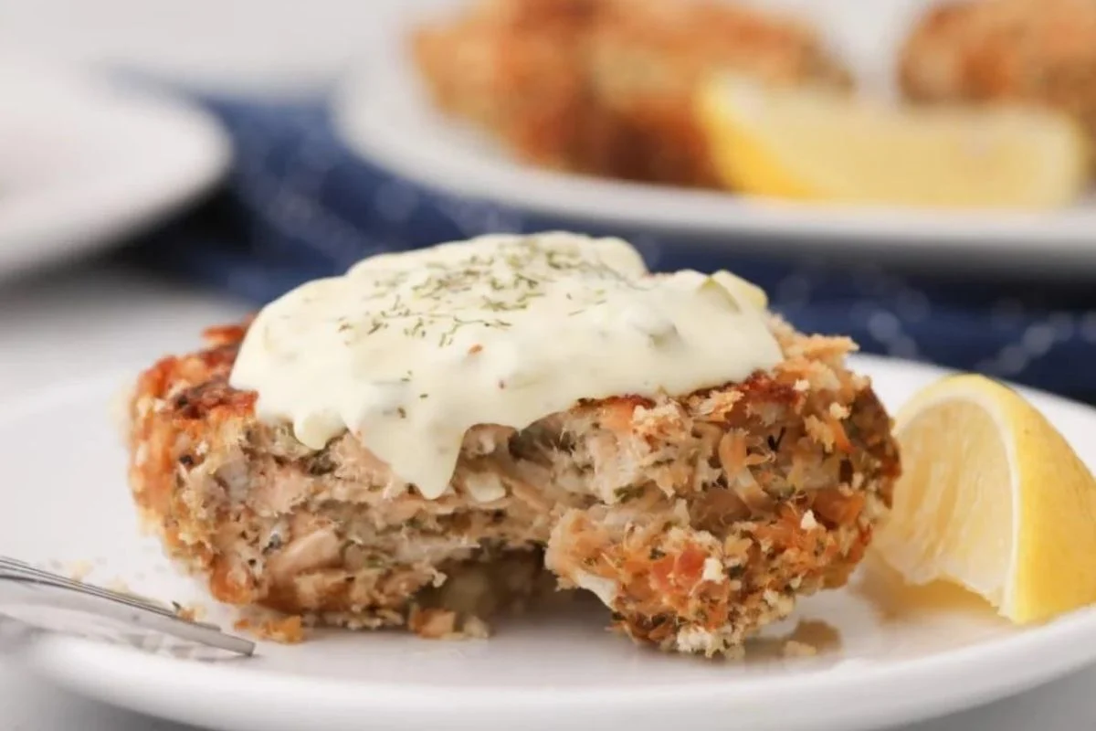A close-up of a salmon patty topped with a creamy sauce and dill, served on a white plate with a lemon wedge, and more patties in the background cooked using a smoker.