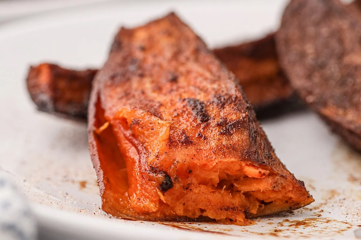Close-up of a roasted sweet potato wedge with a crispy, seasoned exterior and soft interior on a white plate. Perfect as sides for your favorite smoked dinners.